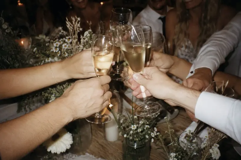 Guests clink champagne glasses over a table of flowers at a wedding.