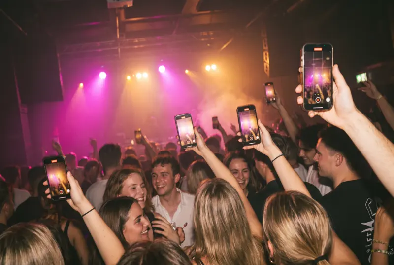 A crowd at a club holds up phones to photograph the stage, under pink and amber lights.