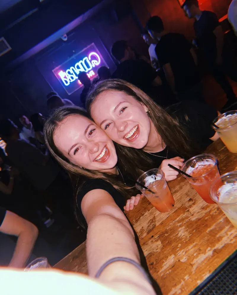 Two friends laugh taking a selfie at the bar, with a purple neon sign behind them.