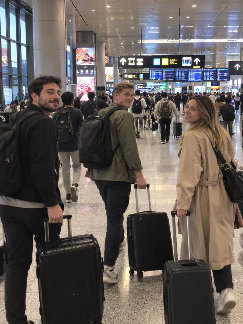 Three friends pull their suitcases through an airport and turn toward the camera.