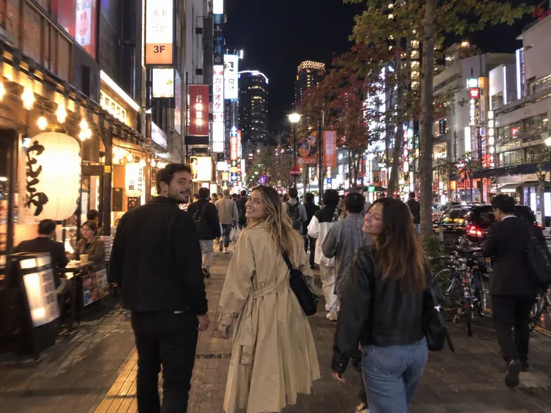 Three friends look back smiling on a neon-lit Tokyo street at night.