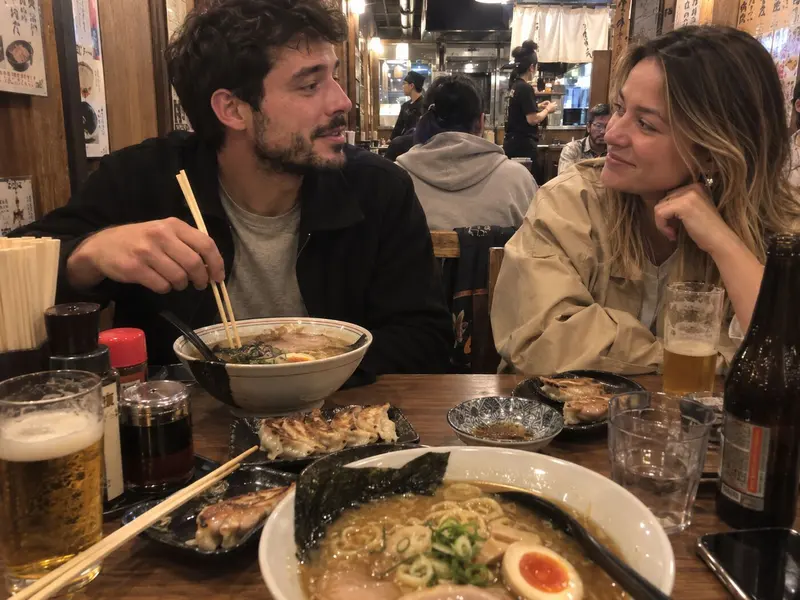 Two friends laugh over bowls of ramen and beers.